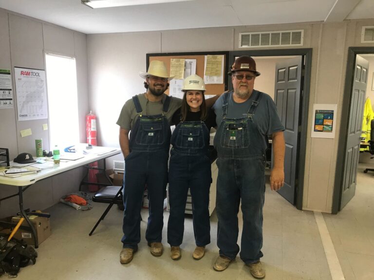 Three people wearing hard hats and overalls standing in an office-like setting.