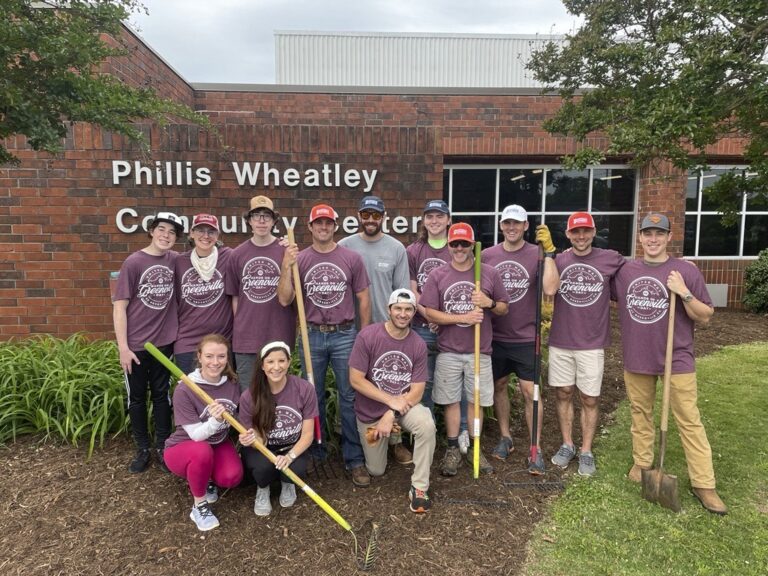 A group of people in matching shirts from Brasfield and Gorrie Greenville pose with gardening tools in front of the Phillis Wheatley Community Center building.