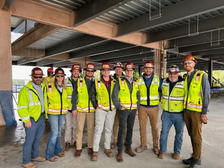 A group of eleven people in construction hard hats and high-visibility safety vests stand inside a Brasfield and Gorrie office building frame in Raleigh.