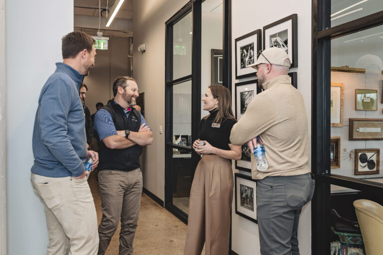 A group of people talk in the hallway of a modern office. Framed pictures are visible on the wall behind them.