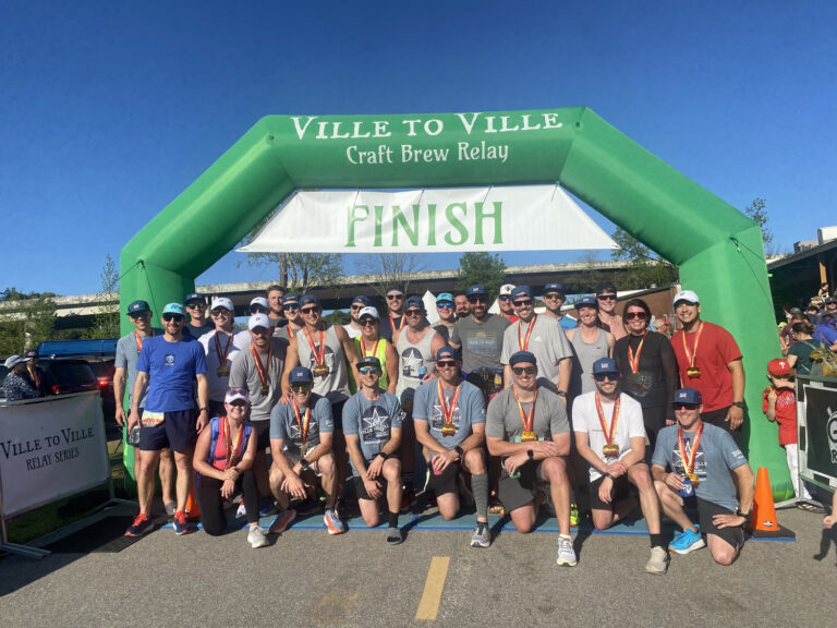 A group of runners wearing medals pose together under a green Ville to Ville Craft Brew Relay finish line arch on a sunny day in Greenville, sponsored by Brasfield and Gorrie.