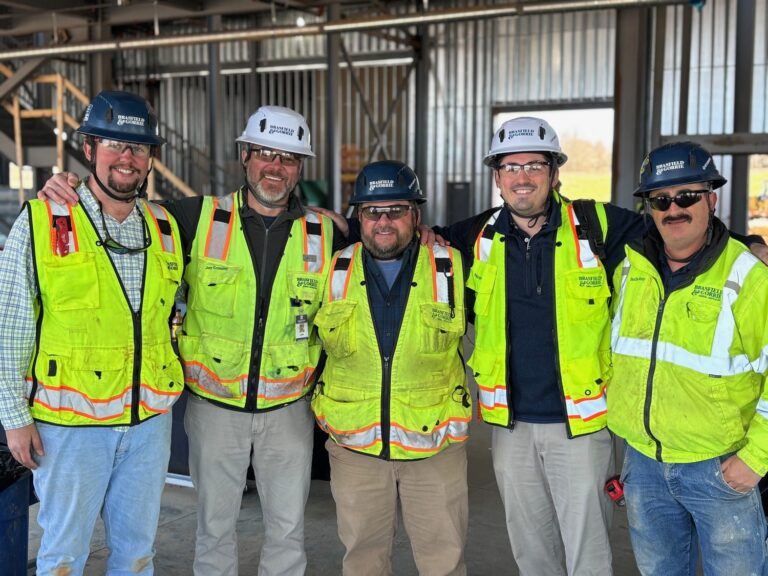 Five construction workers wearing hard hats and high-visibility vests pose for a group photo inside a building under construction by the Huntsville Office of Brasfield and Gorrie.
