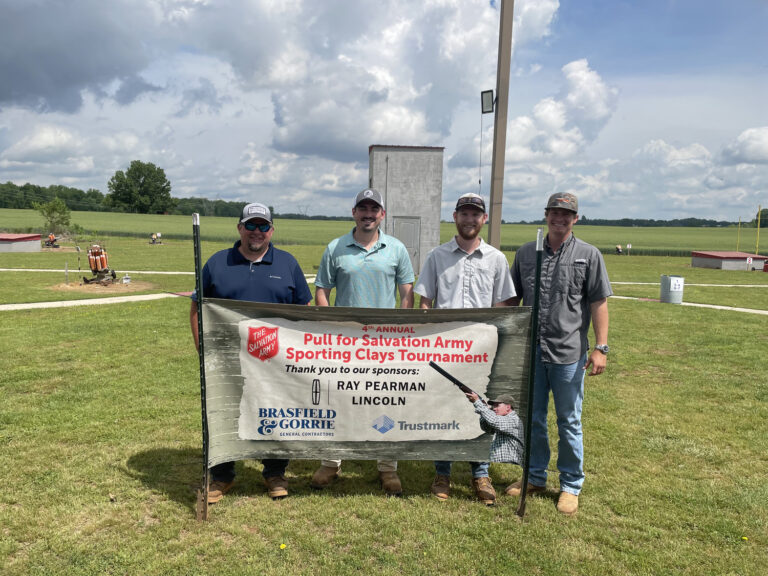 Four men from the Huntsville Office of Brasfield and Gorrie stand outdoors behind a banner for the Salvation Army Sporting Clays Tournament, with grassy fields and a cloudy sky in the background.
