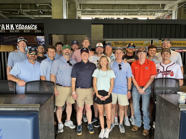 A group from the Columbus Office of Brasfield and Gorrie stand and smile for a photo in front of a concession stand at a sports venue. Most are casually dressed, with some wearing baseball caps and jerseys.