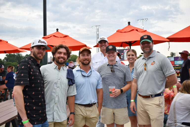 A group of seven people, mostly men, from the Columbus Office Brasfield and Gorrie, pose together outdoors at a baseball stadium near orange umbrellas on a cloudy day.