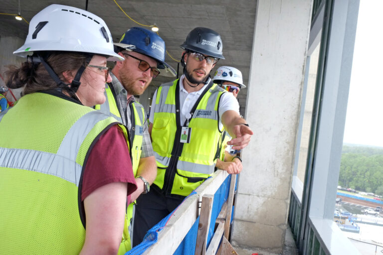 Four construction workers in safety gear stand on a high floor of the Orlando Office, discussing and pointing out details while overlooking a Brasfield and Gorrie construction site through large windows.
