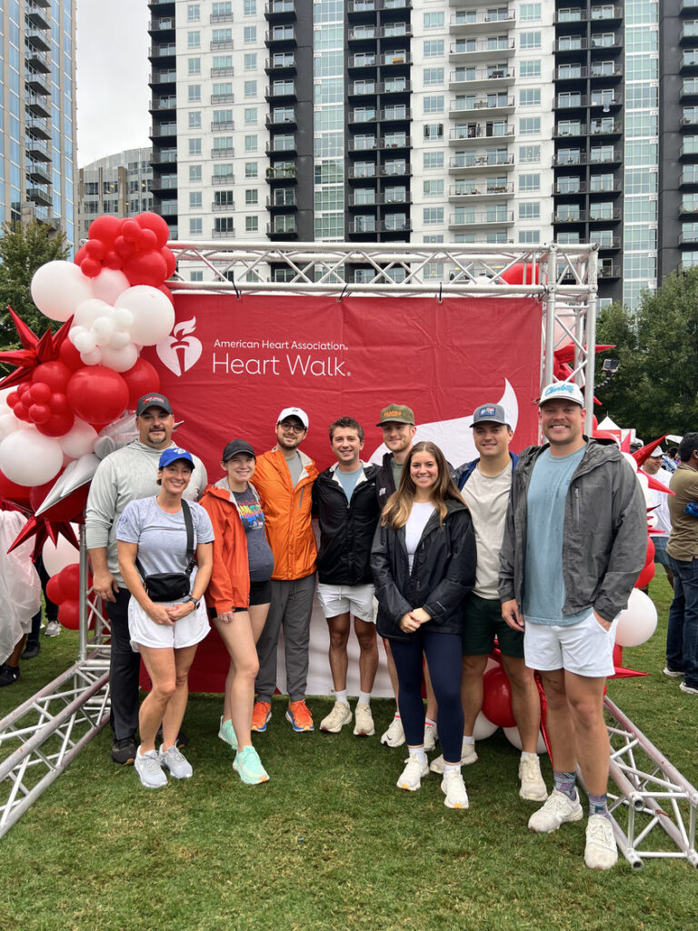 A group of nine people from the Charlotte Office of Brasfield and Gorrie pose in front of a red Heart Walk banner with red and white balloons, outdoors with tall buildings in the background.