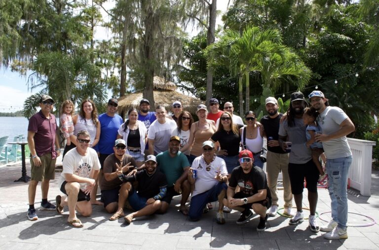 A group from the Orlando Office of Brasfield and Gorrie pose together outdoors by a waterfront, surrounded by palm trees and greenery, under a sunny sky.
