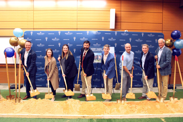 Nine people in business attire from the Raleigh office of Brasfield and Gorrie pose with shovels in a ceremonial groundbreaking for the Perry Family Simulation Hospital.