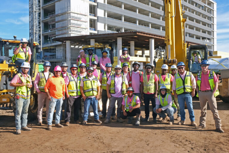 A group of construction workers from the Orlando Office Brasfield and Gorrie, in safety gear, pose for a photo at a work site with machinery and a partially built structure in the background.