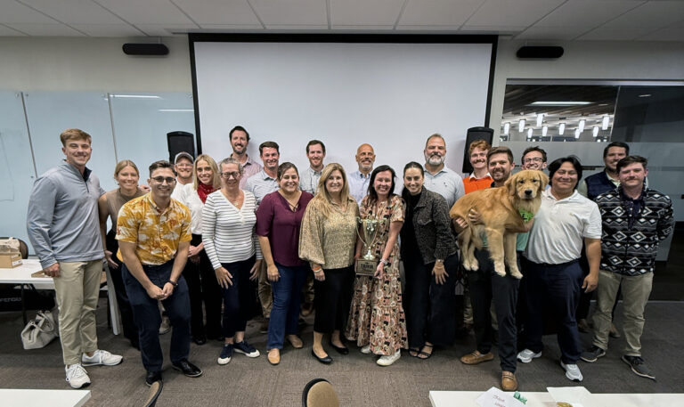 A group of about 25 adults and a golden retriever from the Orlando Office of Brasfield and Gorrie pose together in a conference room in front of a projector screen.