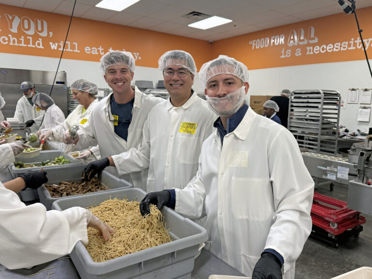 Three people in white lab coats, hairnets, and gloves stand in a commercial kitchen at the Orlando Office of Brasfield and Gorrie, sorting food with others. The background features orange walls adorned with motivational text.
