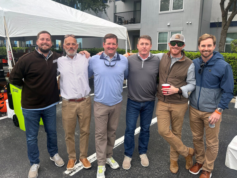 Six men from the Orlando Office of Brasfield and Gorrie stand side by side outdoors, smiling at the camera under a white tent, with buildings and greenery in the background.