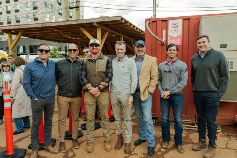 Seven men stand side by side outdoors in front of a red shipping container at what appears to be a construction site for the Greenville Office of Brasfield and Gorrie.