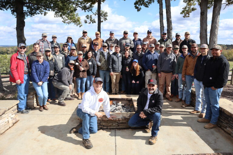 A large group of people from the Charlotte Office of Brasfield and Gorrie pose outdoors around a circular stone fire pit, with trees and a scenic landscape in the background.