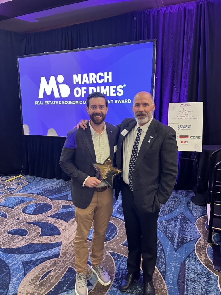 Two men in suits stand together smiling; one holds a star-shaped award. Behind them is a 