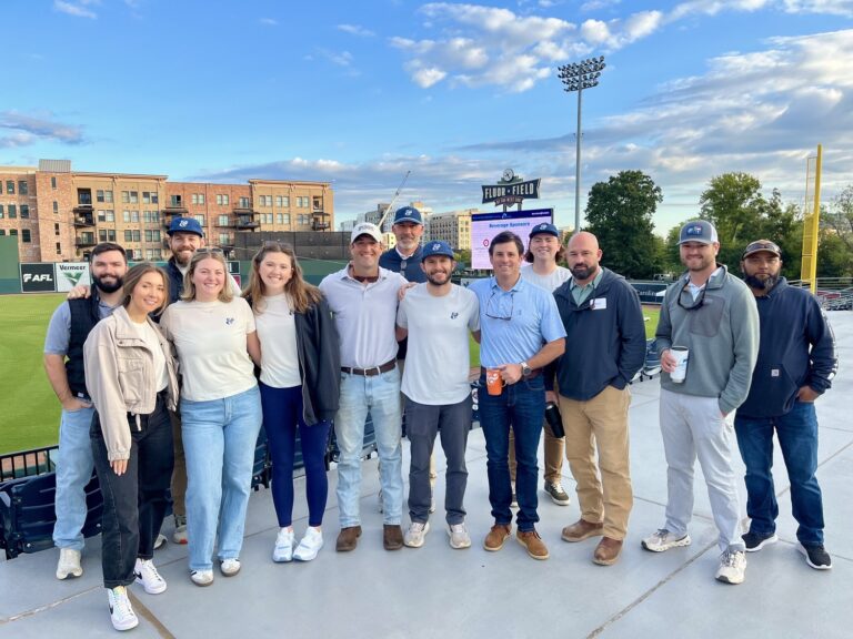 A group of thirteen people poses outdoors on a terrace at a sports stadium, with a grassy field, scoreboard, and buildings visible in the background, celebrating a Brasfield and Gorrie Greenville team event.