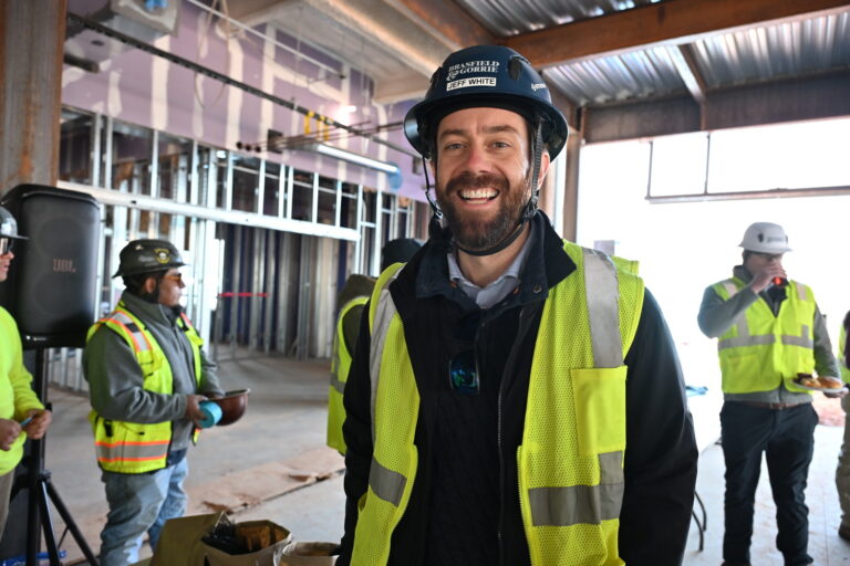 A smiling man in a safety vest and hard hat stands inside a Brasfield and Gorrie Greenville building under construction, with other workers in the background.