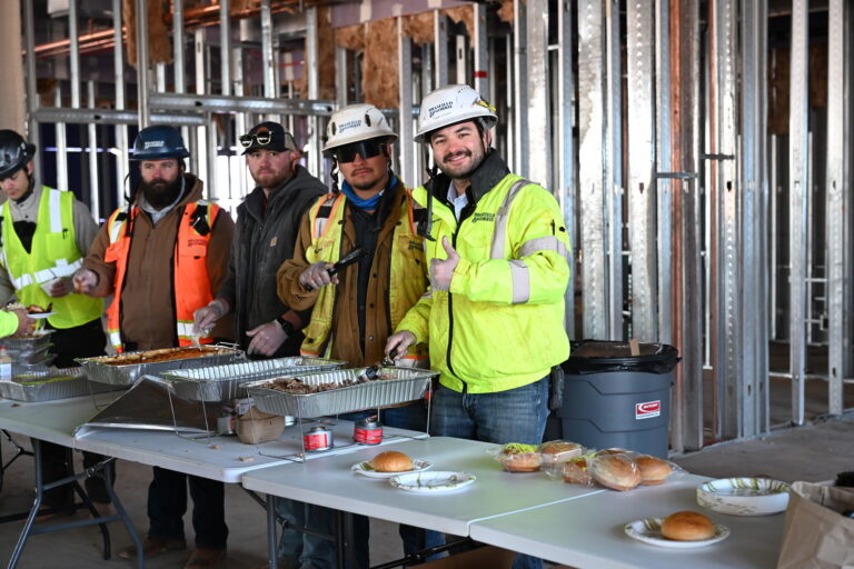 A group of construction workers in safety gear serve themselves food from trays set on tables inside an unfinished Brasfield and Gorrie Greenville building.