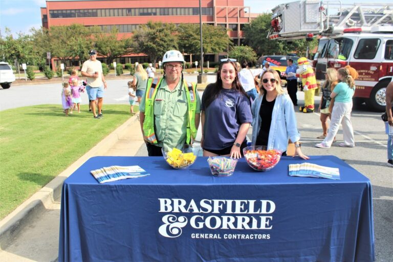 Three people stand behind a table with brochures and candy bowls at a Columbus Office Brasfield and Gorrie event, with a fire truck and people in the background.