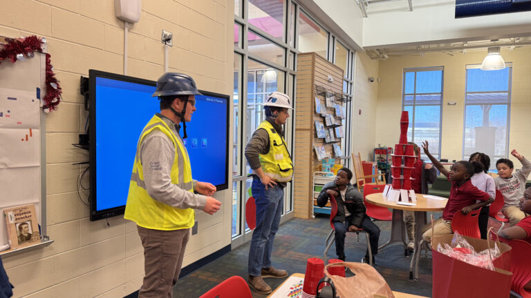 Two people in safety vests and helmets from the Columbus Office of Brasfield and Gorrie stand in a classroom near students, with a stack of red cups on a table and a large screen on the wall.