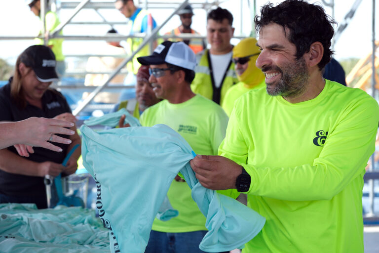 Un hombre con una camiseta de color amarillo neón sonríe mientras recibe una camiseta azul claro en un acto al aire libre organizado por la oficina de Brasfield and Gorrie en Jacksonville, rodeado de otras personas con ropa similar.