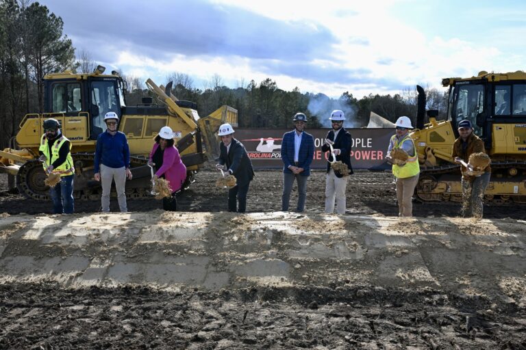 A group of people in construction gear participate in a Brasfield and Gorrie Greenville groundbreaking ceremony with shovels on a dirt site, flanked by two bulldozers.