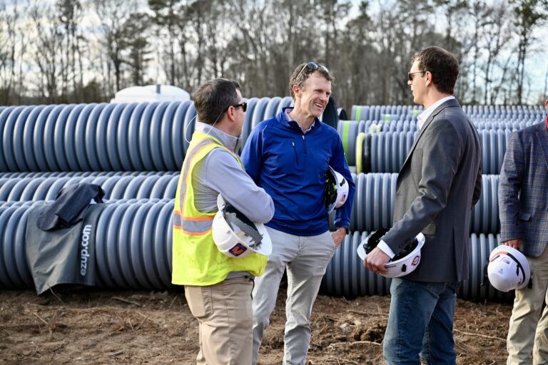 Three men stand and talk at a Brasfield and Gorrie Greenville construction site, holding hard hats, with large stacks of pipes and trees visible in the background.