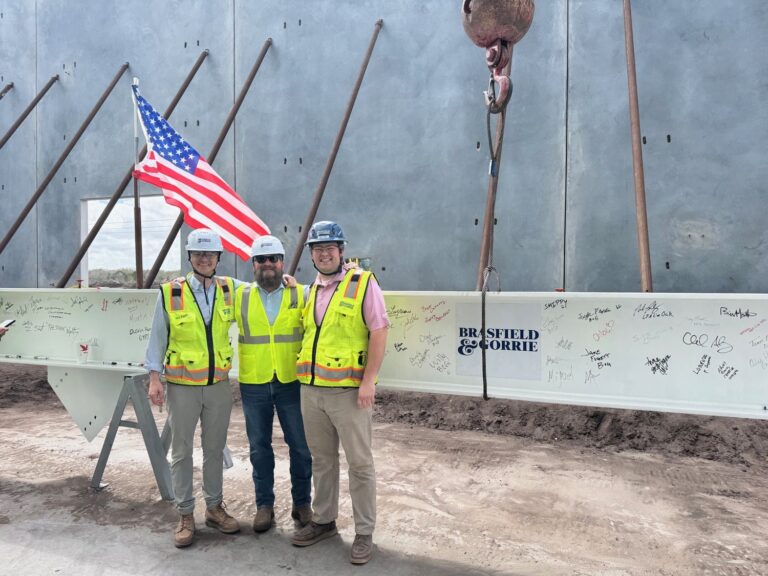 Three people in hard hats and high-visibility vests stand before a signed steel beam at a Brasfield and Gorrie Nashville office site, positioned in front of a concrete wall with a crane hook overhead.