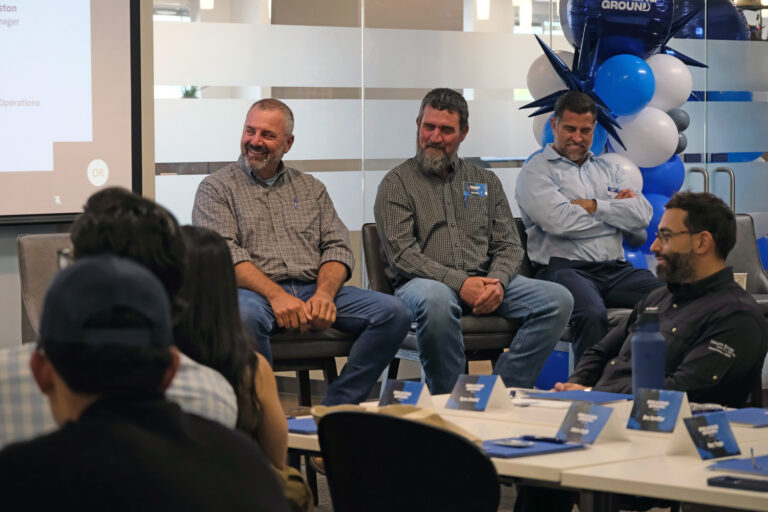 Three men sit at the front of a room addressing an audience in the Orlando Office of Brasfield and Gorrie, with blue and white balloons and name cards visible in a professional setting.