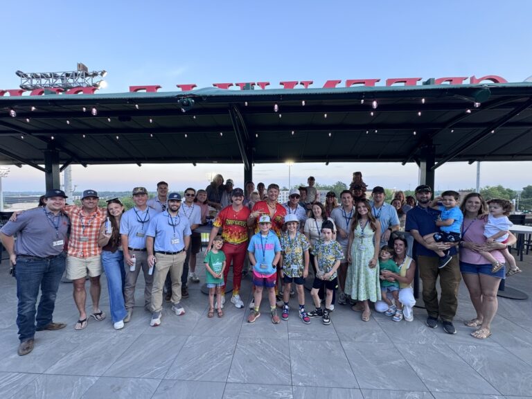 A large group of adults and children, some wearing Brasfield and Gorrie Greenville event lanyards and casual summer clothing, pose together outdoors under a pavilion.