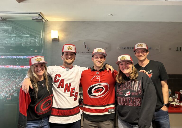 Five people from the Raleigh office of Brasfield and Gorrie wearing Carolina Hurricanes jerseys, apparel, and matching hats posing in a stadium suite.
