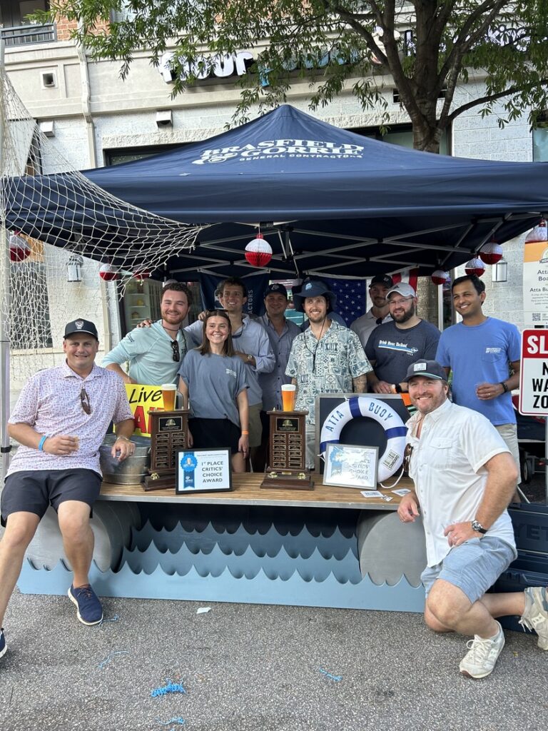 A group of people from the Raleigh office of Brasfield and Gorrie stand around a table featuring two “1st Place Critics’ Choice Award” trophies under a branded canopy.