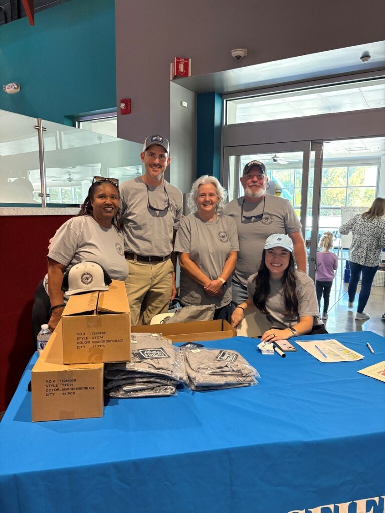 Five people from the Brasfield and Gorrie Raleigh office wear matching grey shirts behind a table of hats and apparel at an event.