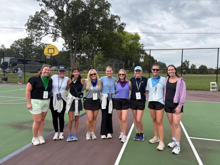 Nine women from the Nashville Brasfield and Gorrie office standing in a line on a pickleball court wearing athletic clothing, neck gaiters, and event badges.