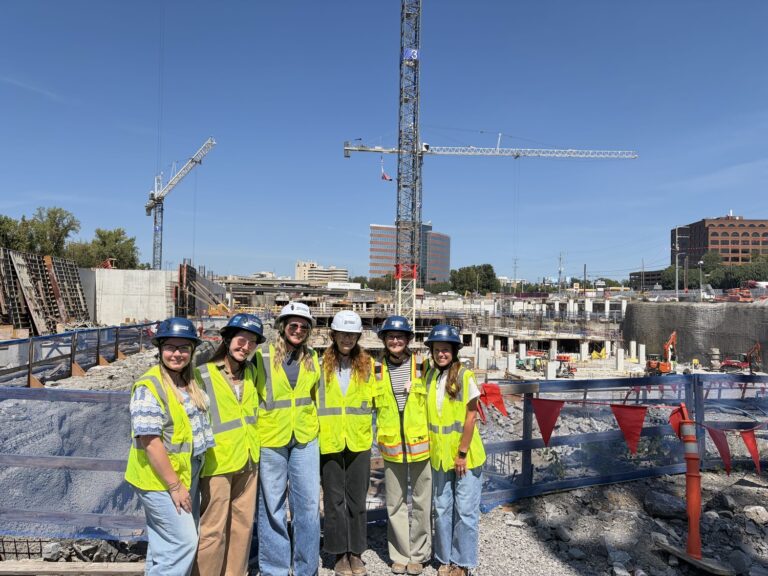 Six people wearing hard hats and high-visibility safety vests stand in a row at a Brasfield and Gorrie construction site in Nashville, with cranes in the background.