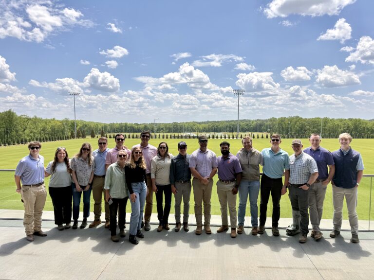 A group of sixteen people from the Atlanta Office of Brasfield and Gorrie stand side by side outdoors on a concrete terrace, with a large green field and trees in the background under a partly cloudy sky.