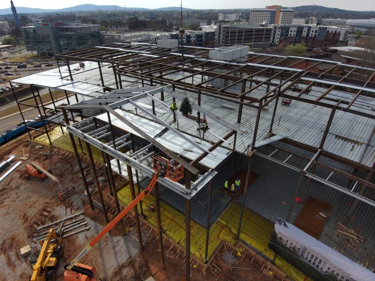 Construction workers install metal decking on a steel framework of a new building against an urban backdrop.