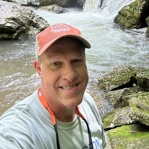 Scott Duckworth, wearing a red cap and light shirt, takes a selfie in front of a rocky stream with flowing water and lush greenery.