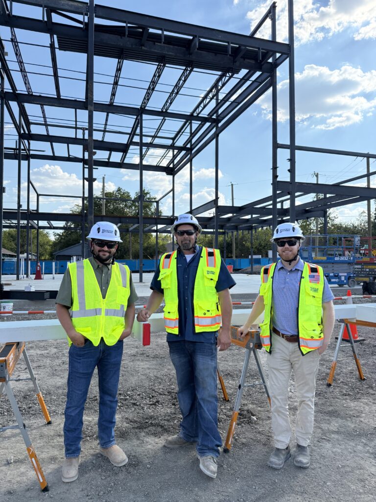 Three men wearing hard hats and high-visibility vests stand at a Dallas Office Brasfield and Gorrie construction site, with a steel building framework in the background.