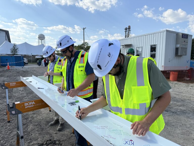 Three construction workers in safety gear sign a steel beam at an outdoor Dallas Office Brasfield and Gorrie construction site, with portable buildings and equipment in the background.