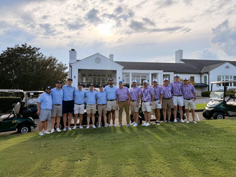 A group of men in golf attire from the Columbus Office Brasfield and Gorrie stand in a row on grass in front of a clubhouse, with golf carts visible on both sides under a partly cloudy sky.