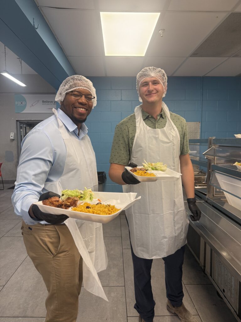 Dos hombres que llevan redecillas para el pelo, guantes y delantales están de pie en una cafetería con bandejas de comida en las manos, sonriendo a la cámara. Esta alegre escena muestra a los voluntarios de la oficina de Miami de Brasfield and Gorrie colaborando con su comunidad.