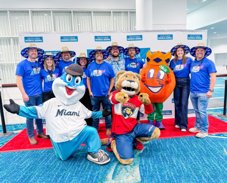 Un grupo de personas con camisetas azules de la oficina de Miami de Brasfield and Gorrie posa con tres mascotas deportivas sobre una alfombra roja frente a un telón de fondo con el logotipo de la empresa.
