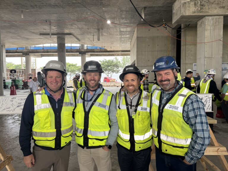Cuatro hombres con chalecos de seguridad y cascos de obra posan en una obra de la sede de Miami, con estructuras de hormigón sin terminar y trabajadores al fondo, lo que da fe del compromiso de Brasfield and Gorrie con la calidad en la construcción.