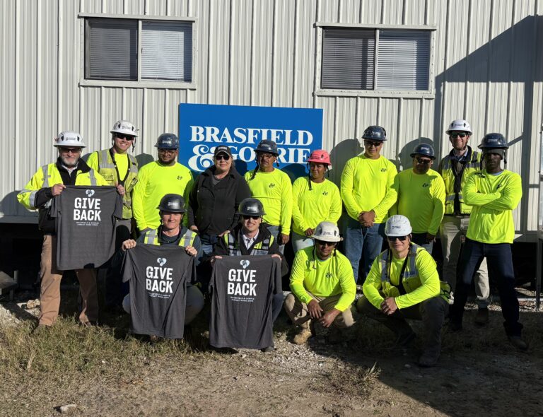 Un grupo de obreros de la construcción, equipados con material de seguridad, se encuentra frente a una caravana con el letrero de Brasfield & Gorrie en la oficina de Jacksonville; dos de ellos sostienen camisetas con el lema «Give Back».