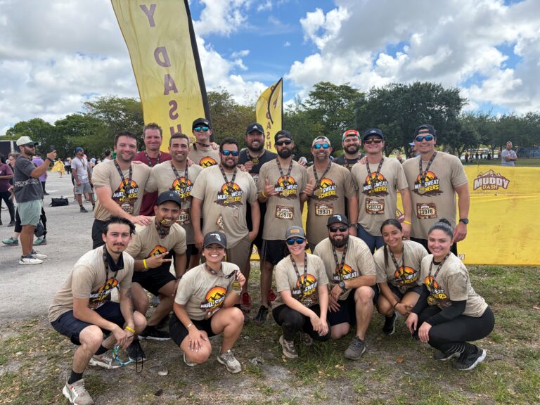 Un grupo de personas de la oficina de Miami de Brasfield and Gorrie, vestidas con camisetas y medallas a juego del Muddy Dash, posan juntas al aire libre delante de pancartas amarillas en un día parcialmente nublado.