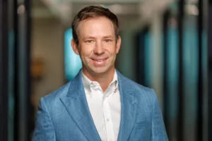Man wearing a light blue blazer and white dress shirt, standing in a hallway with blurred background.
