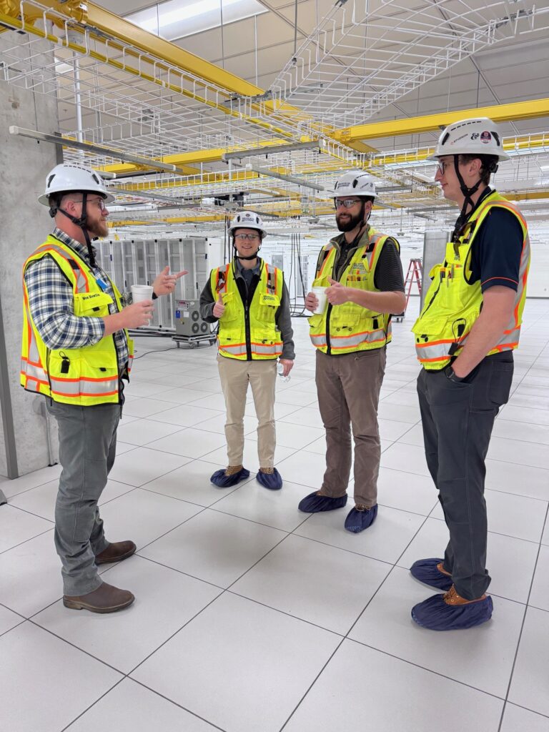 Four construction workers from the Columbus Office of Brasfield and Gorrie, wearing safety vests, hard hats, and shoe covers, stand and talk inside a large industrial facility with exposed cable trays overhead.