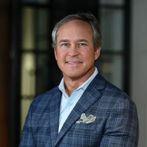 Keith Johnson, dressed in a blue plaid blazer and light pants, sits on a couch, smiling at the camera in a modern indoor setting.